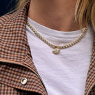 A person wearing a bezel set tennis necklace with a marquise-shaped diamond pendant. The person is also wearing a white t-shirt and a brown check jacket. 