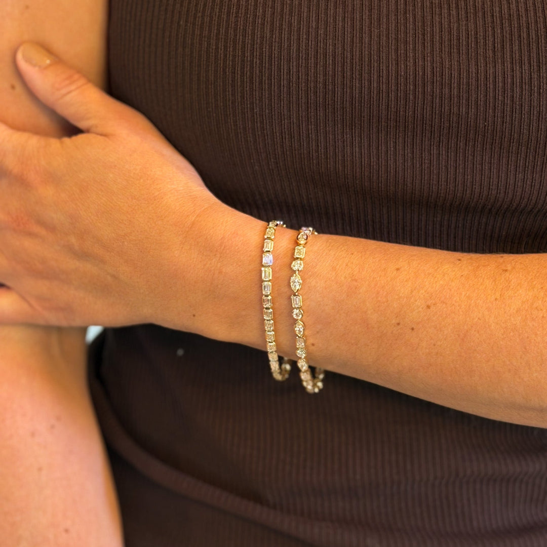 A close-up of a person's wrist wearing two 14k gold tennis bracelets adorned with diamonds, paired with a dark brown ribbed top.