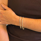 A close-up of a person's wrist wearing two 14k gold tennis bracelets adorned with diamonds, paired with a dark brown ribbed top.