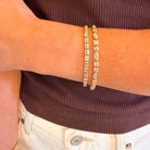A close-up of a person's wrist wearing two 14k gold tennis bracelets adorned with diamonds, paired with a dark brown ribbed top.