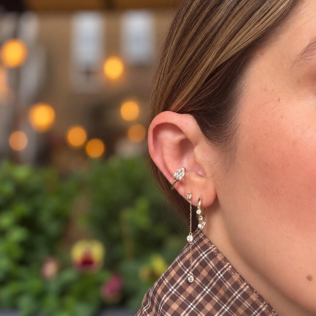 A close-up side profile of a woman wearing three 14k gold earrings with diamond adornments, including a pear-shaped ear cuff. She is wearing a brown and beige plaid shirt against a blurred background with warm lights and green foliage.