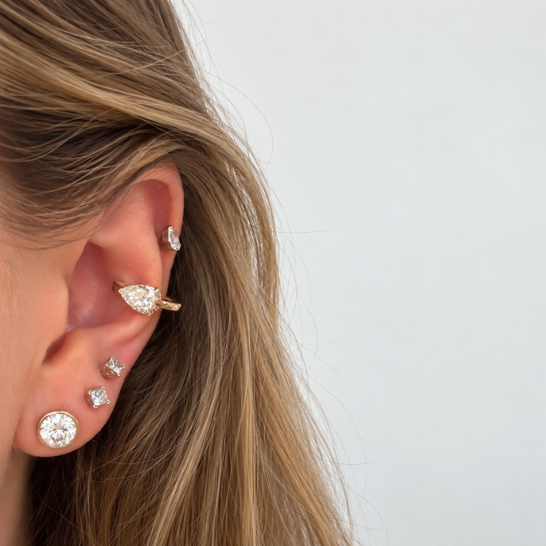 A close-up of a person's ear wearing multiple gold earrings, including a pear shaped ear cuff. 