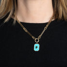 A person wearing a gold chain with a rectangular turquoise, emerald and diamond pendant attached. 