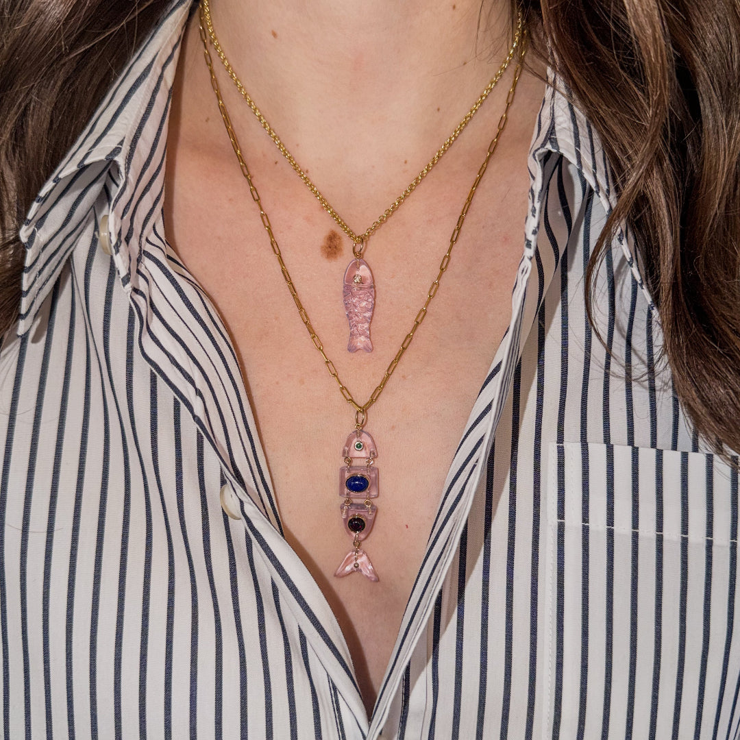 Close-up of a person wearing two 14k gold necklaces with pink quartz fish pendants and a striped shirt.