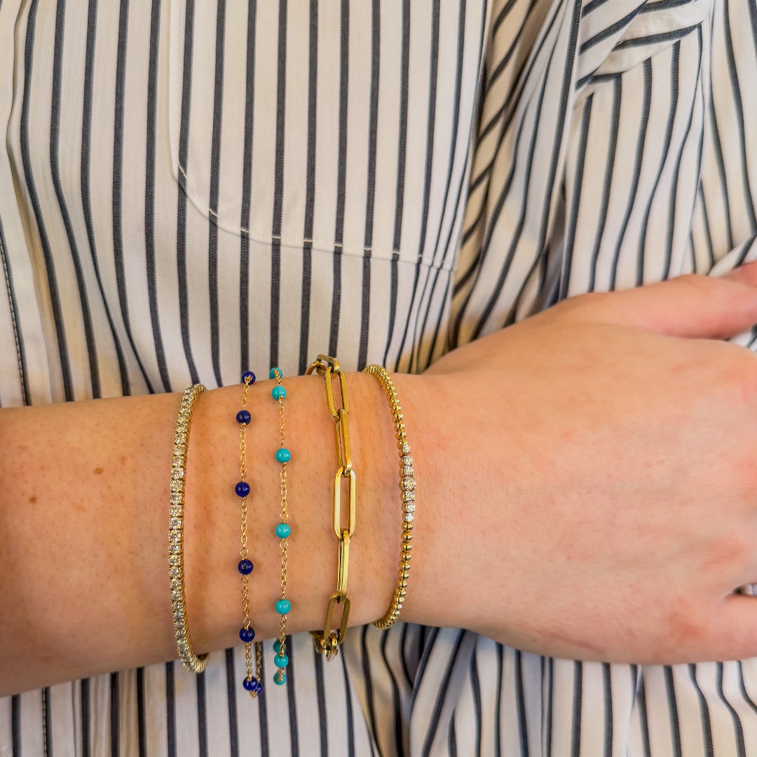 Close-up of a person wearing a striped shirt and her wrist wearing multiple 14k gold bangles with diamonds and beaded stone bracelets with turquoise and lapis.
