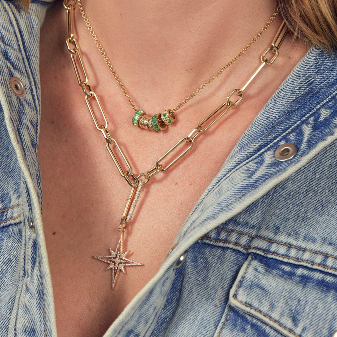 A person wearing a chunky gold chain with a Starburst Diamond Pendant and a thin gold chain with emerald and diamond beads. 