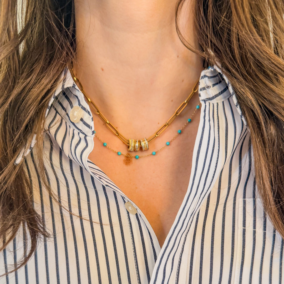 A person wearing a 14k yellow gold turquoise beaded necklace and a 14k yellow gold paperclip chain necklace with beads and a white and black striped button-up shirt. 