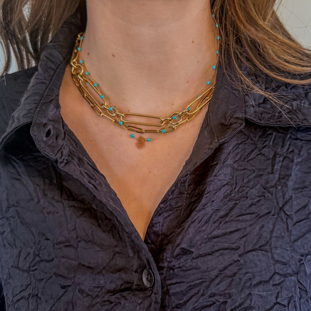 A person wearing a blue shirt and a 14k gold station necklace with turquoise beads layered with two 14k gold chain necklaces. 