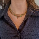 A person wearing a blue shirt and a 14k gold station necklace with turquoise beads layered with two 14k gold chain necklaces. 