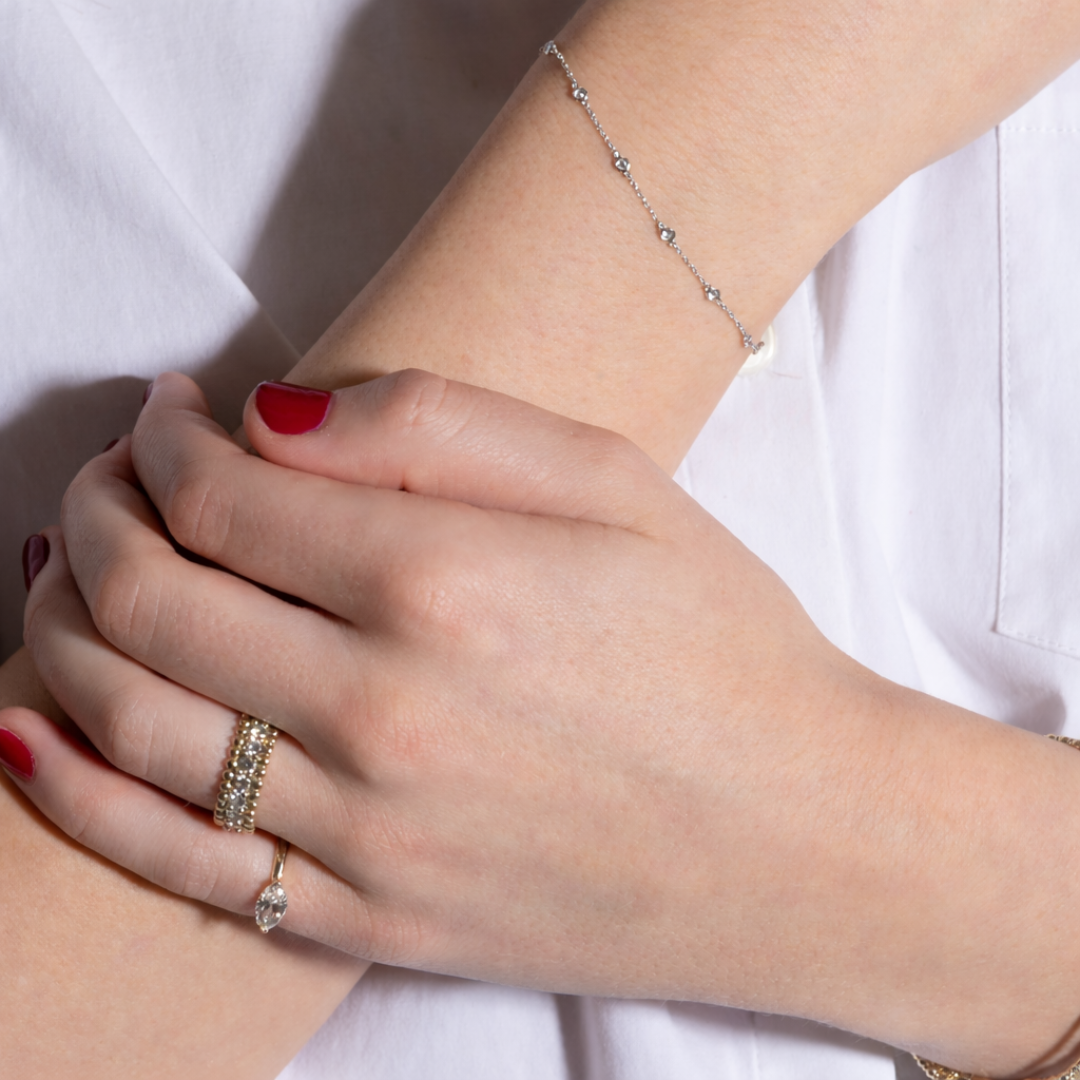 Close-up of a person's wrist with a white gold diamond station bracelet, ring, and nail polish on a white background