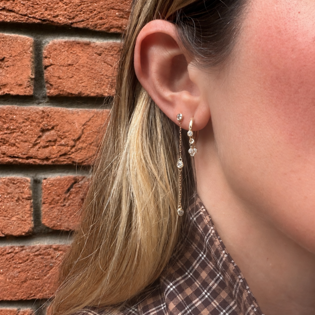A person wearing diamond station drop earrings in a second piercing and diamond drop hoops in the first piercing against a brick wall. 